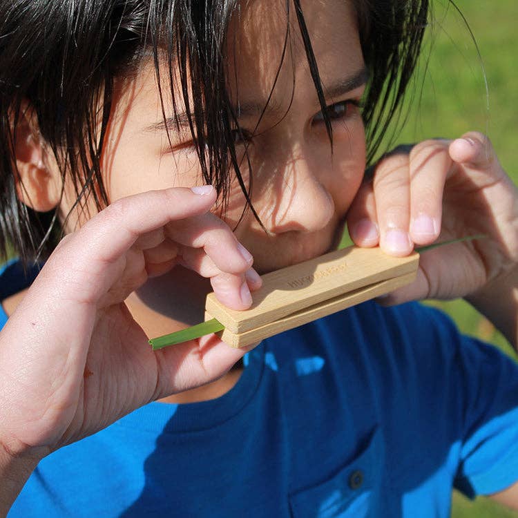 A dark-haired child in a blue shirt plays with the Huckleberry Grass Whistle, holding it to their mouth and blowing on it while enjoying the bright sunlight during an outdoor hike.