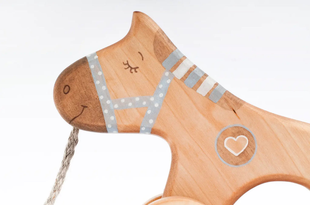 A close-up of a Handmade Wooden Horse Pull Toy | Blue on wheels. The horse is painted with a smiling face and bridle, featuring a heart inside a circle on its body. A string is attached to the front for pulling the toy. The background is plain white.