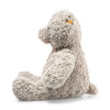 A side view of the Steiff Honey Teddy Bear, 19 inches, from the Soft Cuddly Friends Collection, showcases its curly, textured fur in a light beige hue. It sits upright against a plain white background, displaying a small yellow tag on its ear.