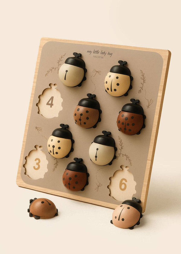 My Little Lady Bug Wooden Puzzle features a wooden peg board with numbered slots and eight ladybug-shaped pieces in assorted brown and beige tones, ideal for counting activities. With one ladybug piece set on the table beside the board, this puzzle offers an engaging Montessori-inspired learning experience.