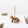 A whimsical hand-painted Red Fox "Party Animal" cake topper shaped like a red fox, with a lit gold candle positioned on its back, surrounded by paper cut-outs of green crowns on a white surface.