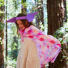 A young girl in a light dress wears Sarah's Silks Magical Creatures Playsilk - Pink Unicorn as a cape and smiles, enjoying imaginative play beneath tall sunlit trees in the forest.