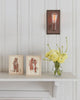 A white mantel displays two framed animal illustrations, a small glass vase of yellow flowers, and the Coral & Tusk Mama Bear and Cub Card—all beneath an exposed bulb sconce on white paneled walls.