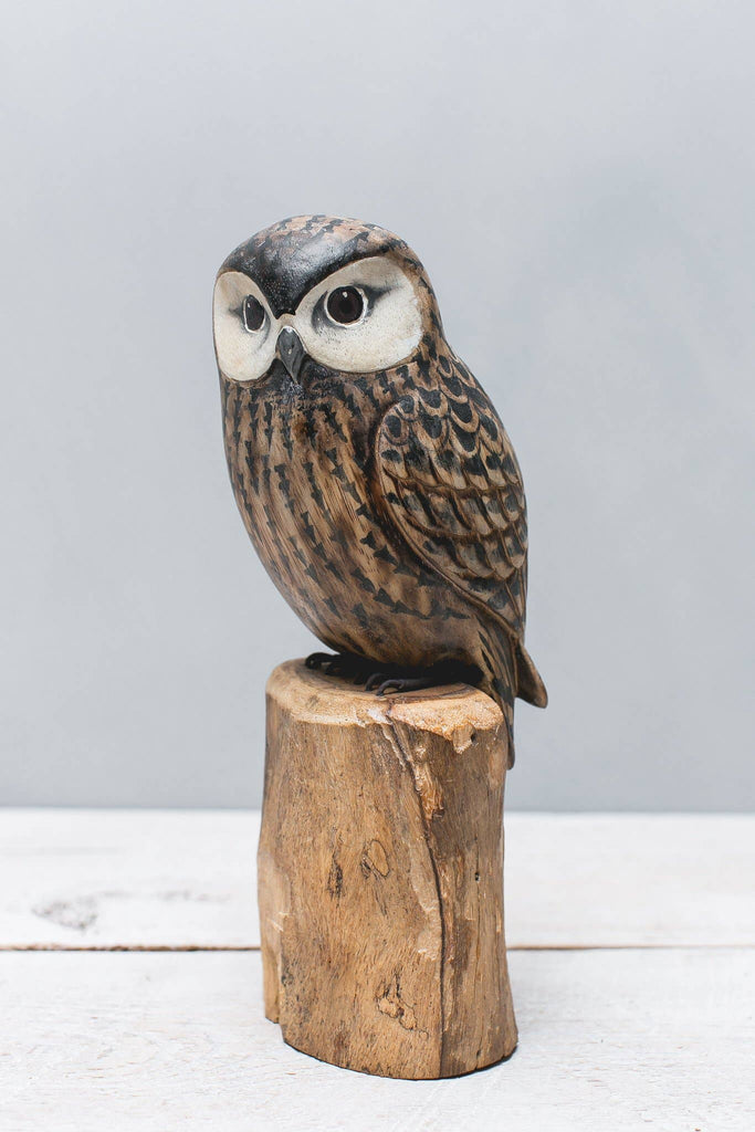 The Hand Carved Ornament - Saw Whet Owl - 10"H is a detailed wooden sculpture with painted feathers, crafted from sustainable softwood, and displayed on a wooden base against a light gray background.