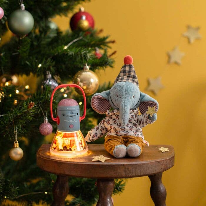 A plush elephant in a party hat sits on a wooden stool next to the Moulin Roty Storybook Lantern - The Minouchkas, with a decorated Christmas tree and yellow star decorations in the background.