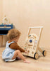 A young child with curly hair, dressed in a brown sweater and light blue dress, sits on the floor playing with the Wooden Activity Walker in a softly lit, minimalistic room.