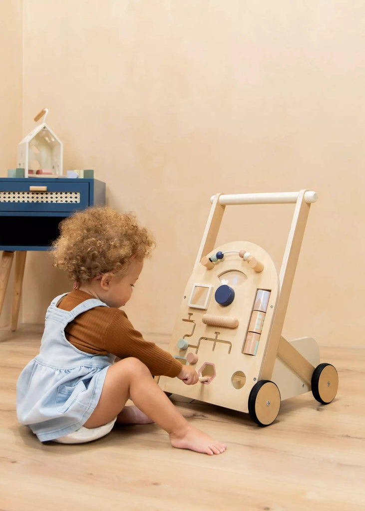 A young child with curly hair, dressed in a brown sweater and light blue dress, sits on the floor playing with the Wooden Activity Walker in a softly lit, minimalistic room.