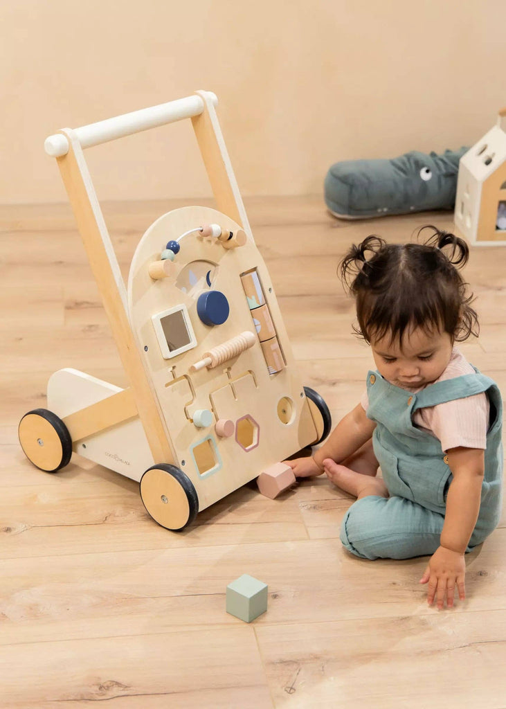 A toddler in blue overalls sits on a wooden floor next to the Wooden Activity Walker, which features colorful blocks and interactive elements. The child looks down, touching the floor and is surrounded by engaging sensory toys that encourage exploration and play.