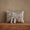 The Coral & Tusk Graveyard Pocket Pillow, featuring an illustrated architectural design and two small fabric figures—a Bunny Ghost and Fox Mummy—rests on a rustic wooden surface against a beige textured wall. Ships in one week.