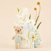 Cuddle + Kind Tiny Charlie the Honey Bear, a hand-knit doll in light blue overalls, stands in front of a floral-themed box with white and yellow flowers and light blue hydrangeas against a beige background.