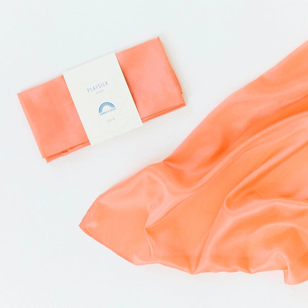 A folded piece of Sarah's Silk Heavy Playsilk Large in peach, labeled "PLAYSILK," lies beside an unfolded one on a light backdrop, ideal for sparking imaginative play.