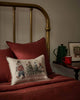 A bed with rust-colored bedding and a brass headboard features the Coral & Tusk Christmas Tree Sled Pillow. On the bedside table are a green coffee pot, a red cup, and a framed picture.