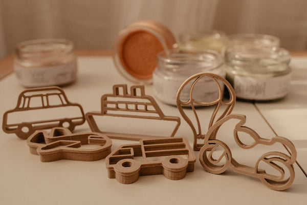 The Transport Cutter Set - Eco Playdough Tool features biodegradable PLA cutters shaped like a car, ship, hot air balloon, airplane, truck, and motorcycle, displayed on a table with glass jars in the background—perfect for creative play as unique vehicle-shaped tools.