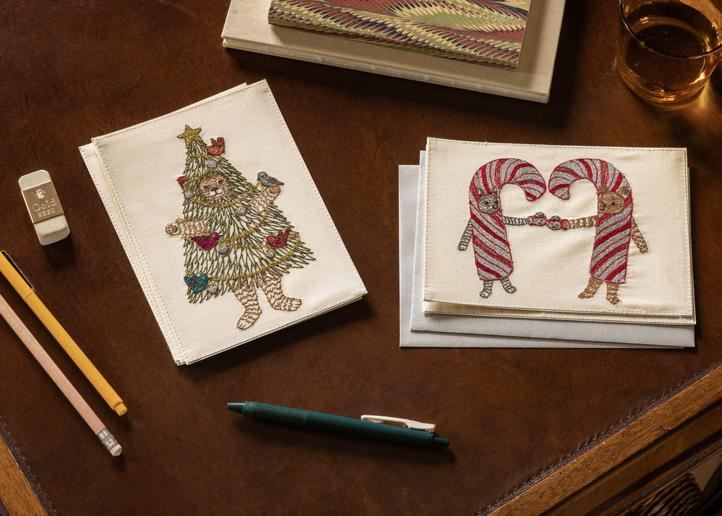 Two embroidered cotton cards on a desk: a Coral & Tusk Christmas Tree Bear Card and another featuring two bears in candy cane costumes holding hands.