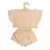 The Mrs. Ertha Doll Clothes - 13″-14″ - Blush Comfy Set is a petite beige doll outfit featuring a short-sleeve crop top and drawstring shorts, made from soft organic cotton, perfectly sized for Minikane dolls and presented on a cardboard hanger.