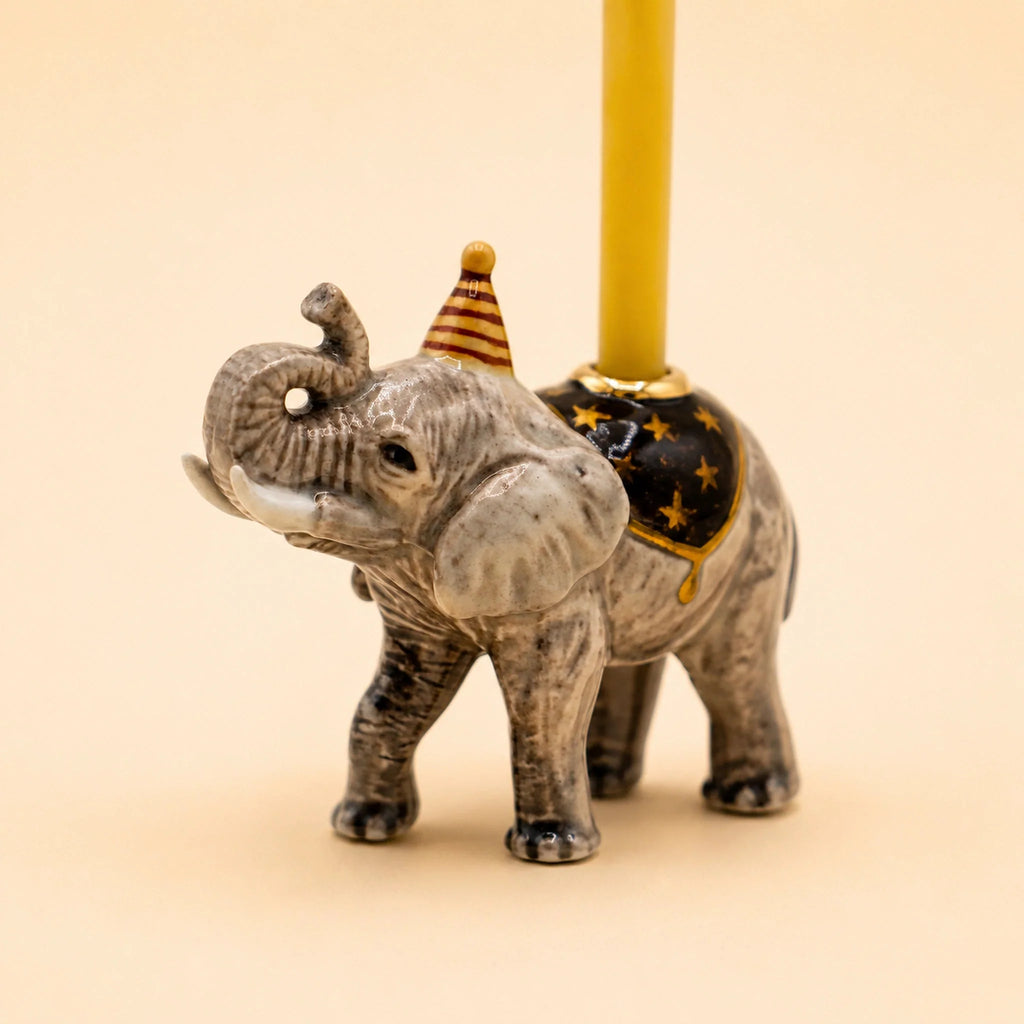 The Elephant Cake Topper - Final Sale (ships in one week) features a hand-painted ceramic elephant in a party hat and gold-star saddle, holding a yellow candle on its back, set against a light beige background.