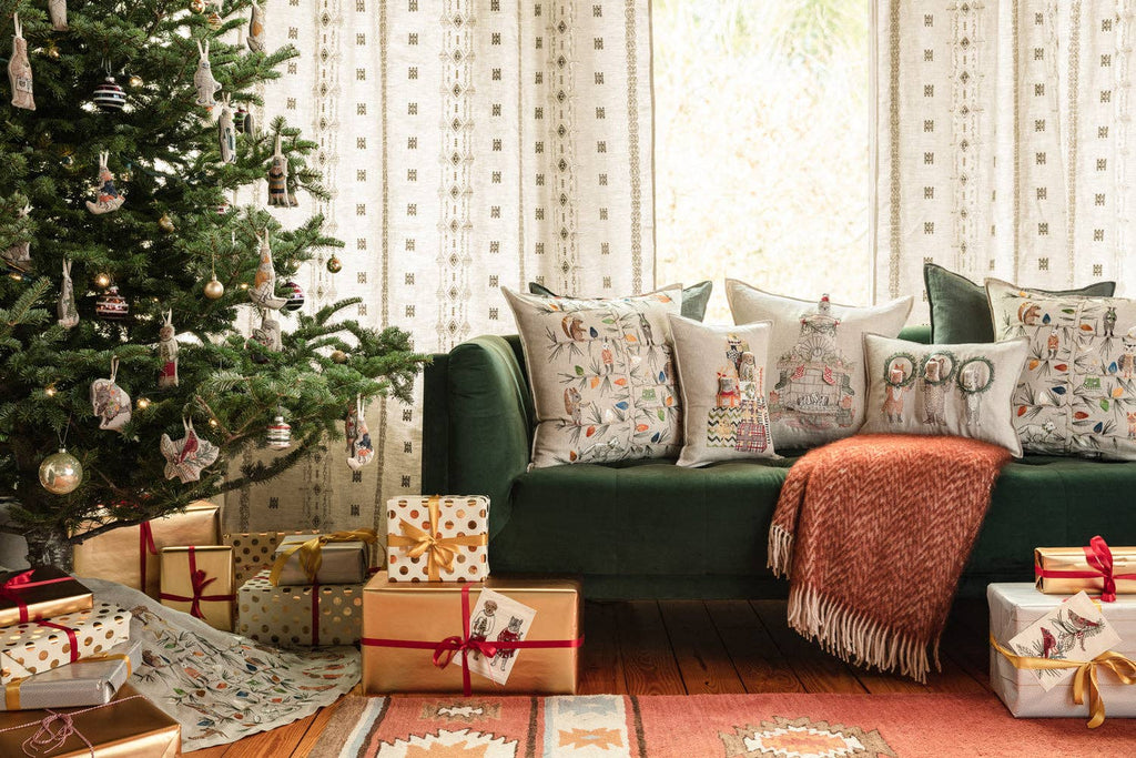 A decorated Christmas tree stands by a green sofa with festive pillows, including the Christmas Wreath Trio Pocket Pillow. Gold and white wrapped gifts sit under the tree, creating a cozy, cheerful holiday atmosphere.