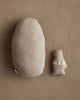 The Coral & Tusk Pocket Easter Egg, an oval-shaped beige fabric pillow, rests on a neutral brown surface beside a small bear-shaped plush labeled "Cozy NUK"—charming additions to any Easter basket.