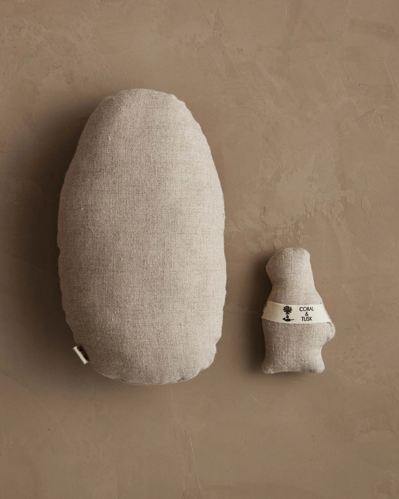 The Coral & Tusk Pocket Easter Egg, an oval-shaped beige fabric pillow, rests on a neutral brown surface beside a small bear-shaped plush labeled "Cozy NUK"—charming additions to any Easter basket.