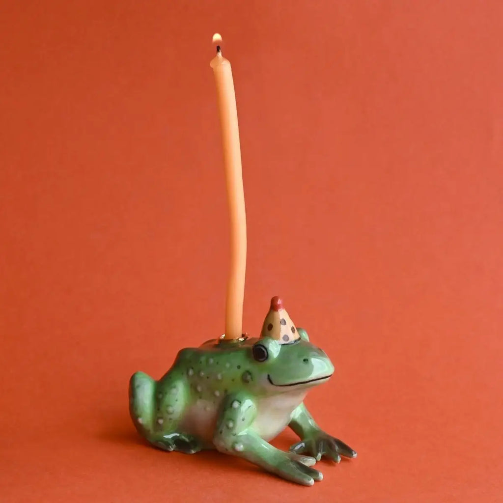 A Frog Cake Topper holding a tall, lit yellow candle on its head, positioned against a solid, vibrant orange background. The frog features a cheerful expression and wears a tiny red party hat.