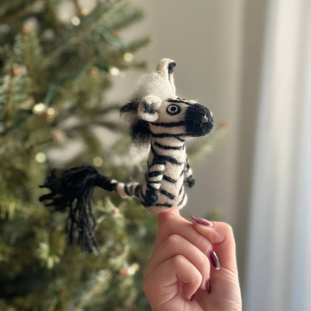 A person holds up the Zebra Finger Puppet in front of a decorated Christmas tree, with natural light streaming through a window behind them.