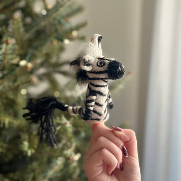 A person holds up the Zebra Finger Puppet in front of a decorated Christmas tree, with natural light streaming through a window behind them.