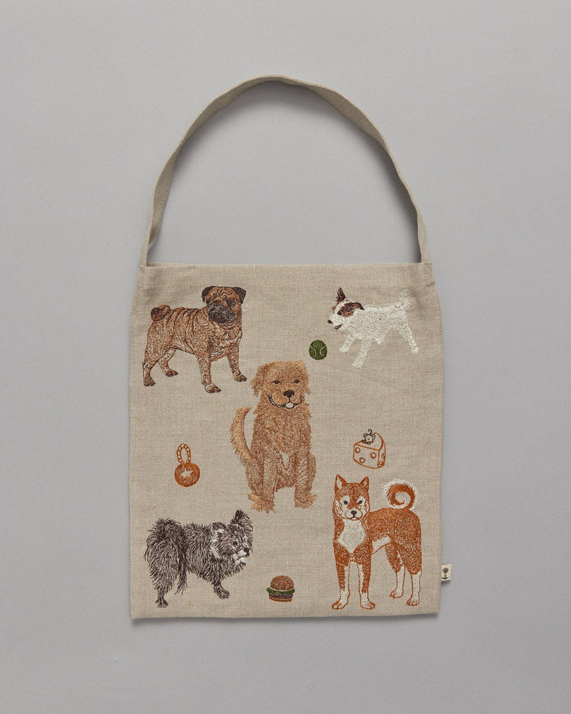 The Dogs and Toys Tote is a playful beige bag with embroidered illustrations of six dog breeds surrounded by objects like balls and chew toys. It features a single strap and is shown against a plain gray background.