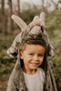 A young boy with blue eyes smiles at the camera outdoors, wearing the Costume - Triceratops Dino hood in gray faux fur, with blurred greenery in the background.