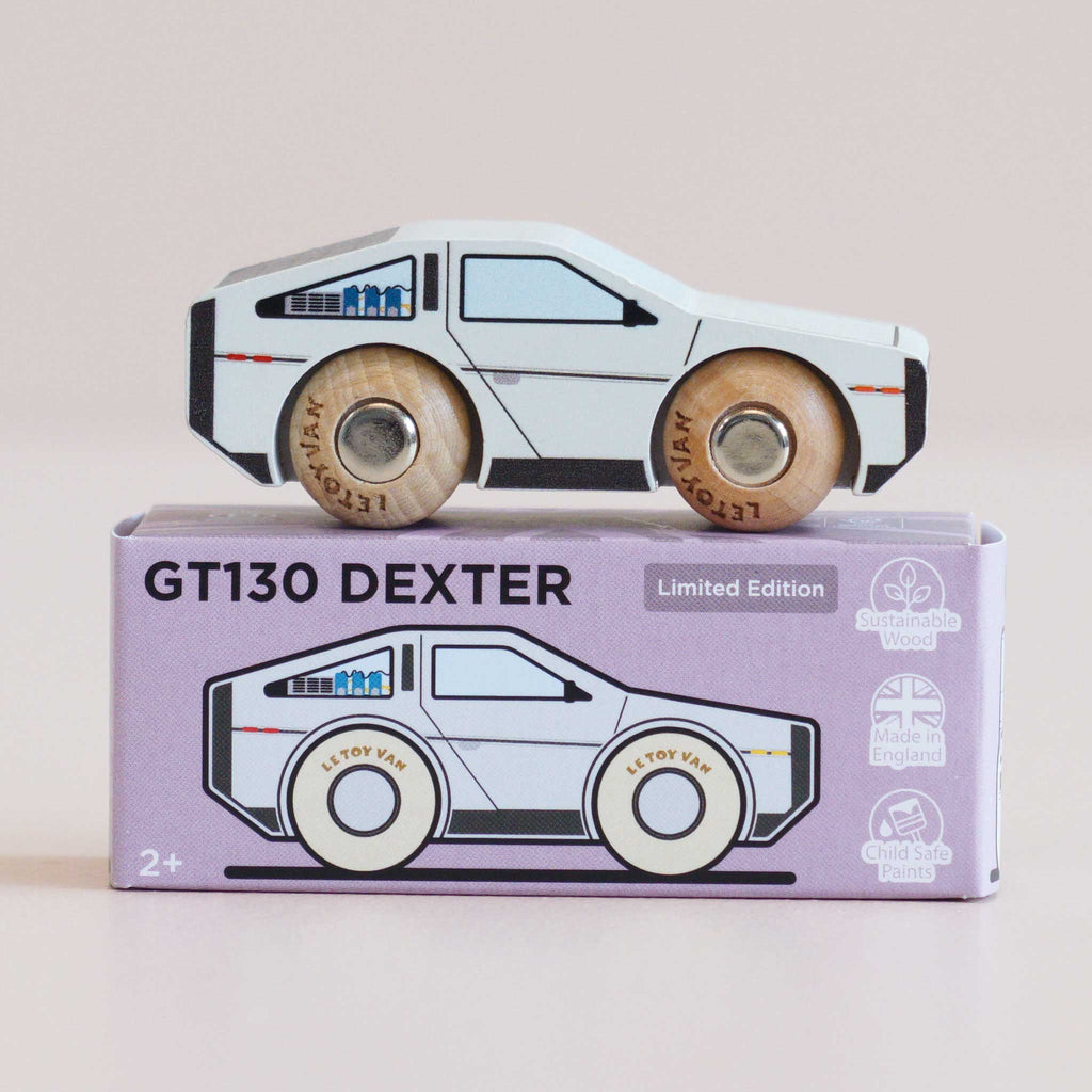 A wooden toy car modeled after a vintage sports car rests atop a box labeled “No. 23 GT130 Dexter | Pre order Aug 2026.” The packaging emphasizes its sustainable wood construction, child-safe paints, and British craftsmanship, making this modern toy ideal for imaginative play.