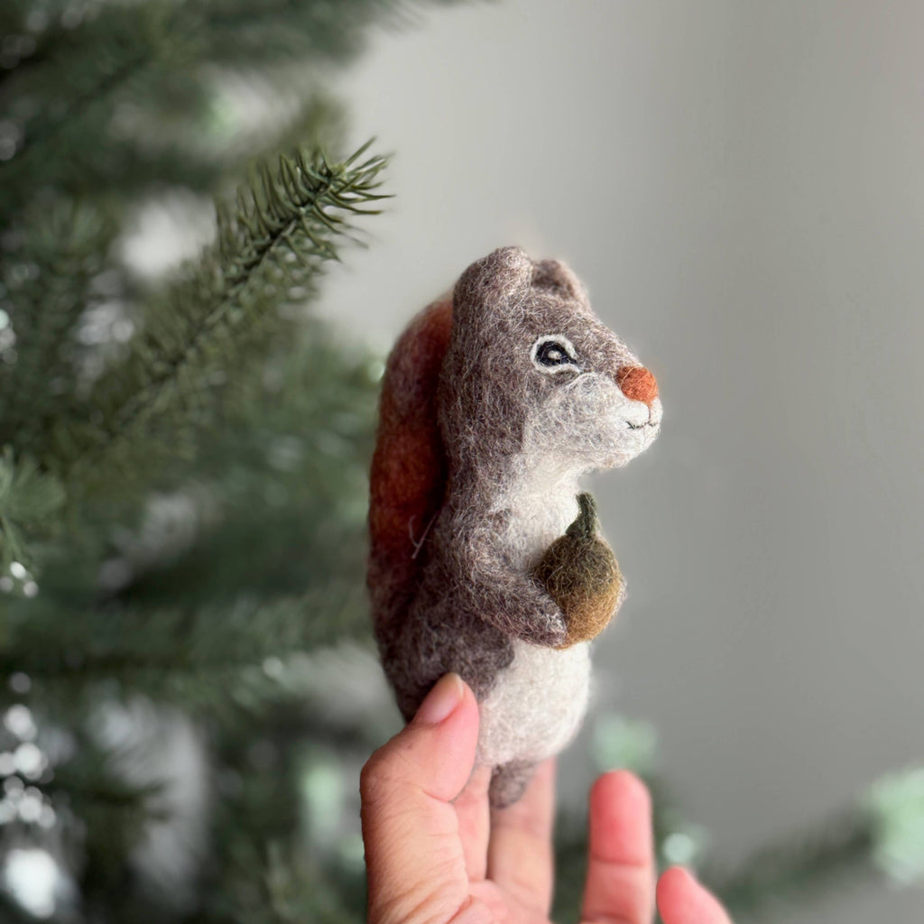 A hand holds the Felt Finger Puppet: Gray Squirrel, featuring a small green acorn in its paws beside pine branches—a cute woodland-themed gift for kids.