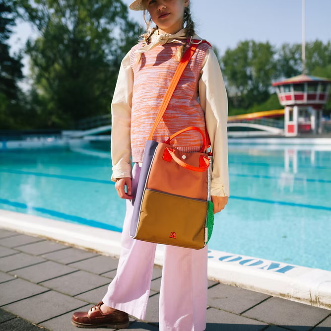 A young girl stands by a pool, wearing a hat, orange sweater vest, light shirt, white pants, brown shoes, and carrying the Sticky Lemon Shoulder Bag | Leather Ball + Golden Cup with orange straps. Trees and a red-and-white building are behind her.