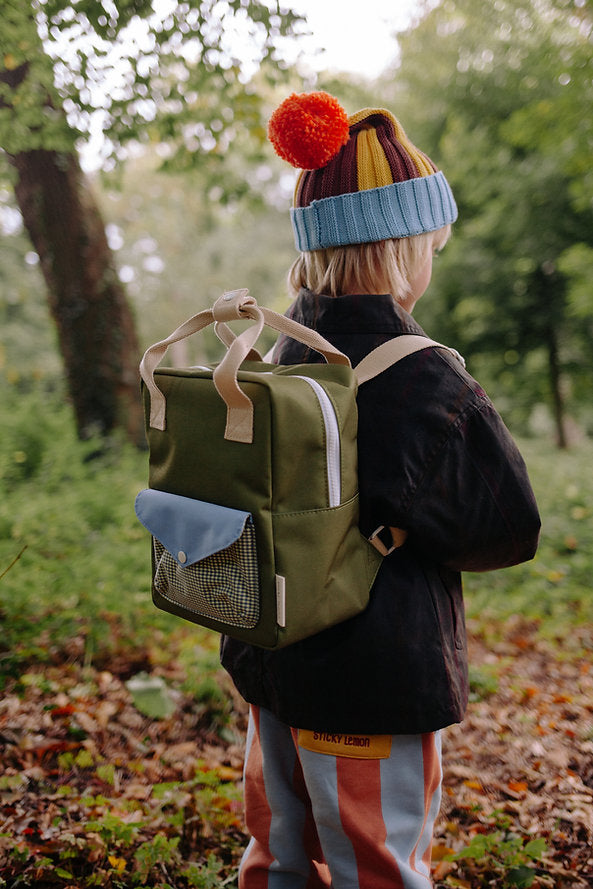 A young child wearing a colorful beanie, striped pants, and a dark jacket stands in a forest with the Sticky Lemon Backpack Small - Envelope - Tickling Grass, made from recycled PET bottles, gazing at the trees.