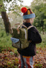 A young child wearing a colorful beanie, striped pants, and a dark jacket stands in a forest with the Sticky Lemon Backpack Small - Envelope - Tickling Grass, made from recycled PET bottles, gazing at the trees.