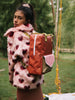 A young girl in a colorful faux fur coat with a Sticky Lemon Backpack Large from the Envelope Collection in Lighthouse Red made of waterproof nylon looks over her shoulder in a lush green park setting.
