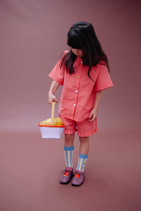 A young girl with long dark hair in a red checkered outfit and colorful socks stands against a brown background, looking down at a Sticky Lemon Treasure Bag - Dewy Lilac filled with yellow plastic fruit.