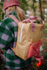 A person in a green and purple checkered fleece jacket and pink beanie carries the Sticky Lemon Backpack Medium - Envelope - Apple Grub, made from recycled PET bottles with pink straps and a waterproof lining, while walking in a forest.