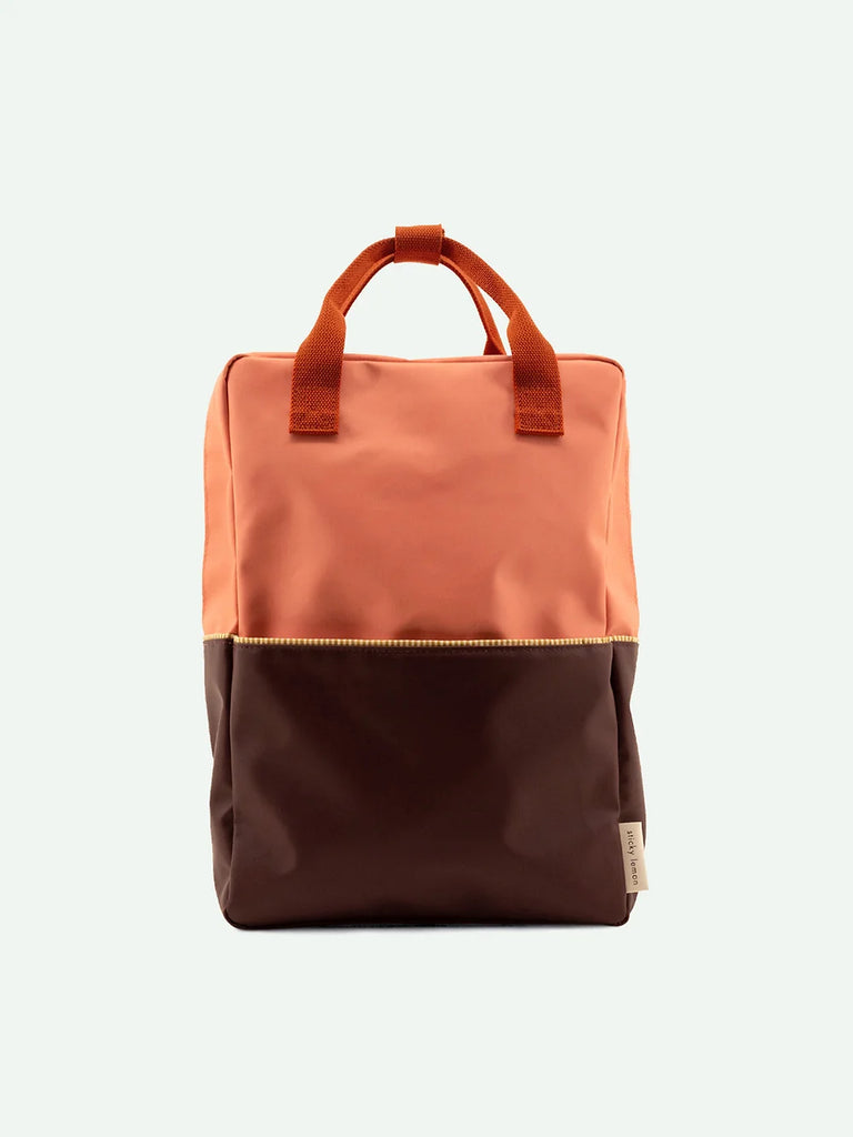 A Meet Me In The Meadows backpack made from recycled PET bottles, with a light orange upper and dark brown lower section, featuring a top handle and a visible front YKK zipper, against a plain background.