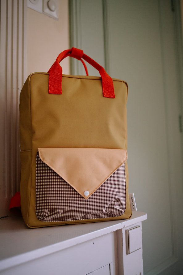 The Sticky Lemon Backpack Large - Envelope - Bookworm Green is a stylish and eco-friendly kids backpack crafted from recycled PET bottles. This waterproof bag features a cheerful yellow color with red handles and a front pocket accented by a beige flap and checked fabric, displayed on a white surface indoors.