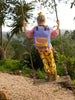A child with a Sticky Lemon Backpack Large made of recycled RPET swings on a rope swing in a lush garden, facing away from the camera. She wears yellow pants with banana prints and brown boots.