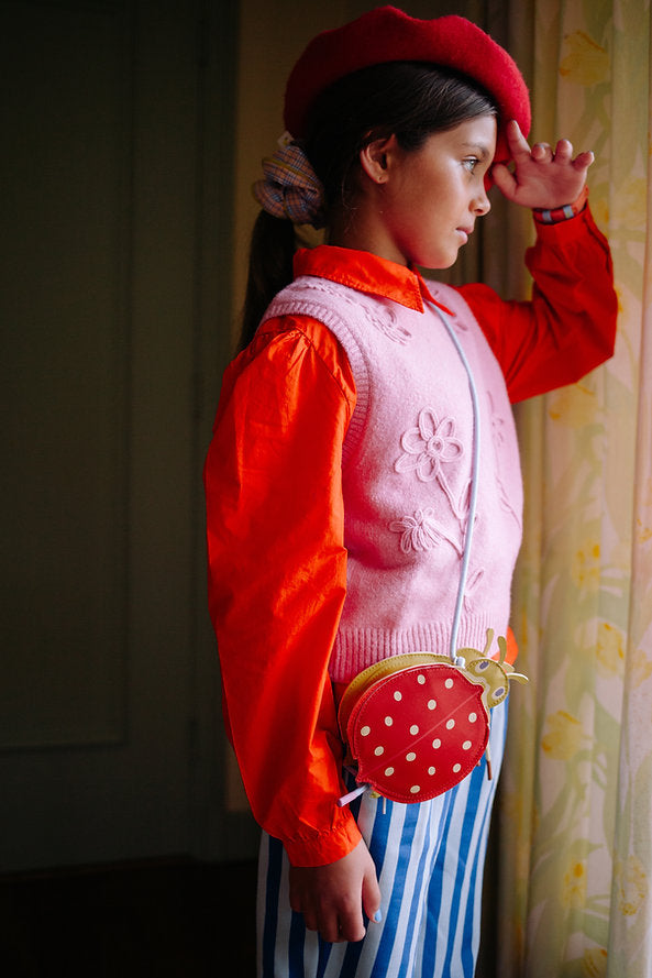 A young girl in a red beret, orange shirt, pink floral vest, and blue-and-white striped pants stands by a window with her hand to her forehead. She carries a charming Sticky Lemon Wallet Bag shaped like a ladybug.
