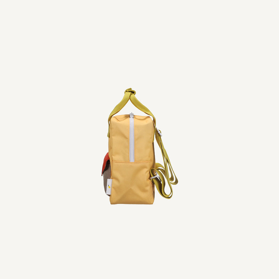 A Sticky Lemon Backpack Small - Envelope - Apple Juice is shown from the side, featuring a yellow rectangular design with a white zipper, yellow handle and strap, plus small red and black accents. The waterproof kids bag is set against a plain white background.