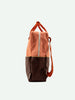 An orange and brown Sticky Lemon backpack with a YKK zipper stands upright against a solid off-white background, featuring adjustable straps and a sleek design.