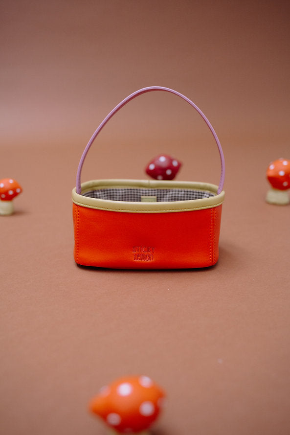 The Sticky Lemon Treasure Bag - Campfire Orange, a small rectangular bag with a pink handle and beige trim, rests on a brown surface surrounded by red-and-white spotted decorative mushrooms.