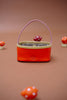 The Sticky Lemon Treasure Bag - Campfire Orange, a small rectangular bag with a pink handle and beige trim, rests on a brown surface surrounded by red-and-white spotted decorative mushrooms.