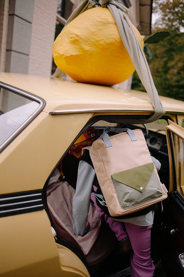 A person carrying a Sticky Lemon Backpack Large - Envelope - Twiggy Beige, possibly made from recycled PET bottles, gets into a yellow car with a large paper mache lemon strapped to the roof by fabric straps.