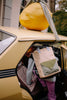 A person carrying a Sticky Lemon Backpack Large - Envelope - Twiggy Beige, possibly made from recycled PET bottles, gets into a yellow car with a large paper mache lemon strapped to the roof by fabric straps.