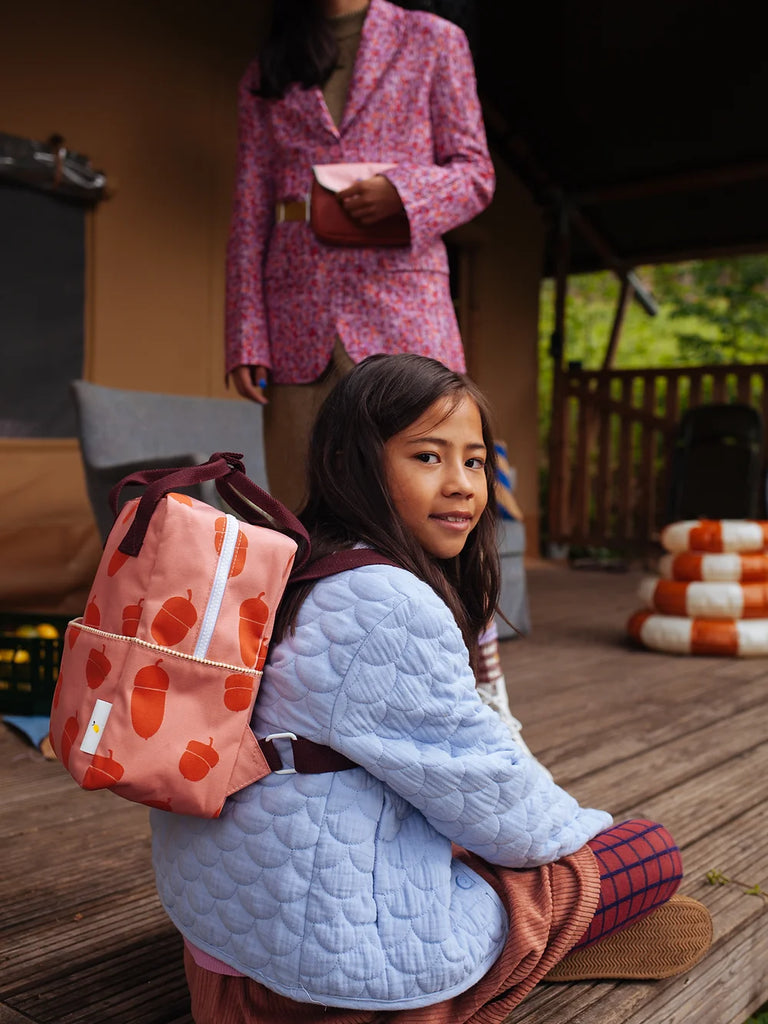A young girl sits on a porch with a Sticky Lemon Backpack Small from the Envelope Collection | Special Edition Acorn, looking over her shoulder. An adult, holding a book, stands in the background, partially out of focus.