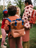 A child wearing a Sticky Lemon Backpack Small in Meet Me In The Meadows color blocking with a YKK zipper stands among other children in a forest-like setting, focusing on the backpack with other children partially visible in the background.