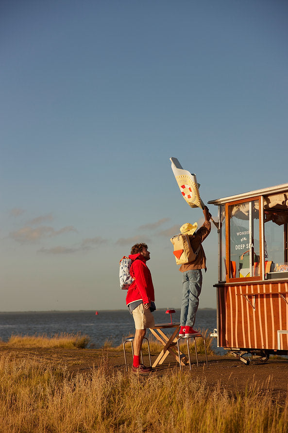 Two people with backpacks—one carrying the Sticky Lemon Backpack Large - Envelope in Reed Green with adjustable shoulder straps—stand by a small wooden food stand near the water. One stands on a stool, placing a large fish sign above the stand, while the other watches. The scene is set on grassy ground under a clear blue sky.