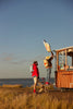 Two people with backpacks—one carrying the Sticky Lemon Backpack Large - Envelope in Reed Green with adjustable shoulder straps—stand by a small wooden food stand near the water. One stands on a stool, placing a large fish sign above the stand, while the other watches. The scene is set on grassy ground under a clear blue sky.